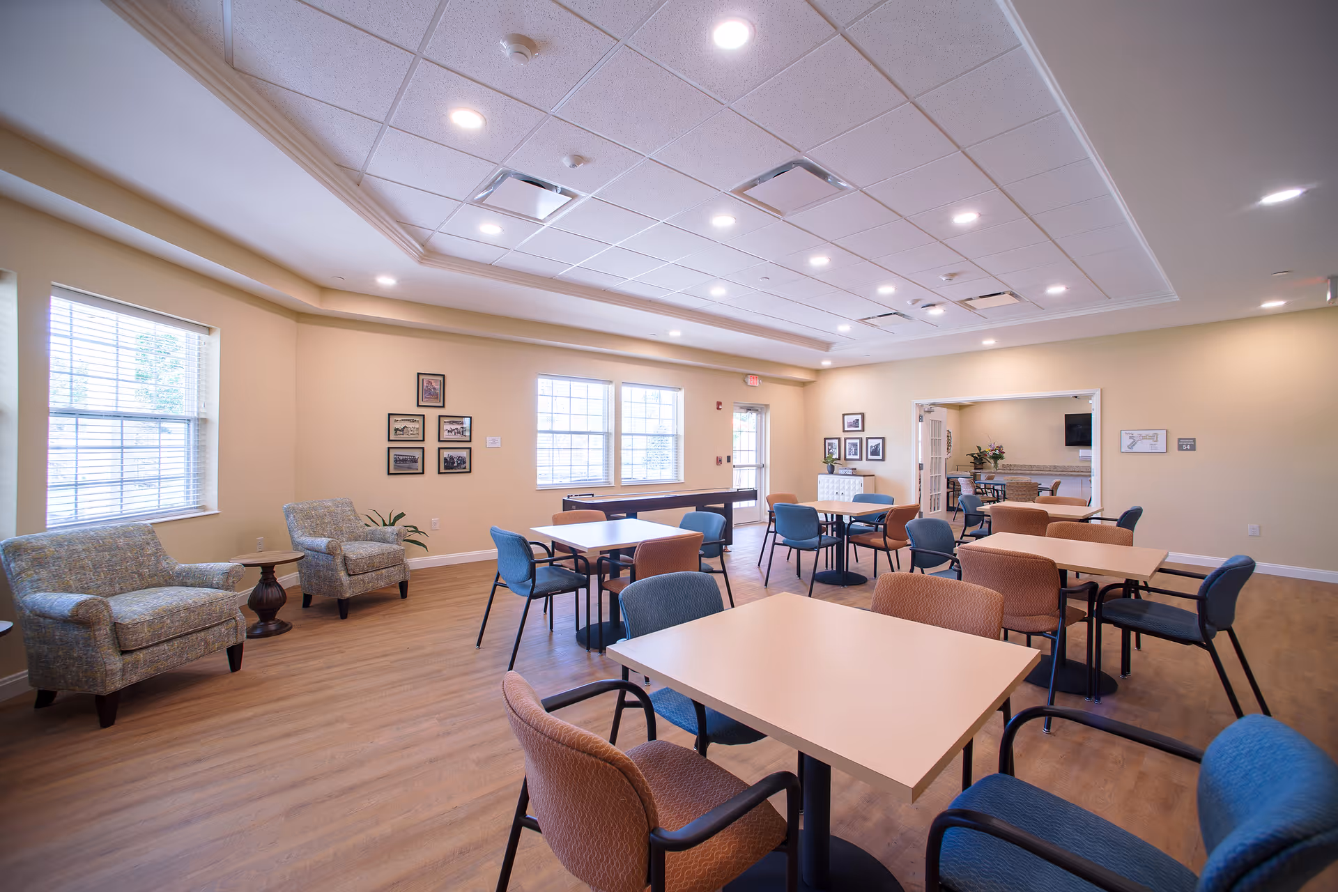 A spacious, well-lit common area in a senior living facility with multiple tables and chairs arranged for group seating. The room has large windows letting in natural light, beige walls, and wood flooring. There are two upholstered armchairs with a small round table near the windows, framed pictures on the walls, and a doorway leading to another room with additional seating and a TV.
