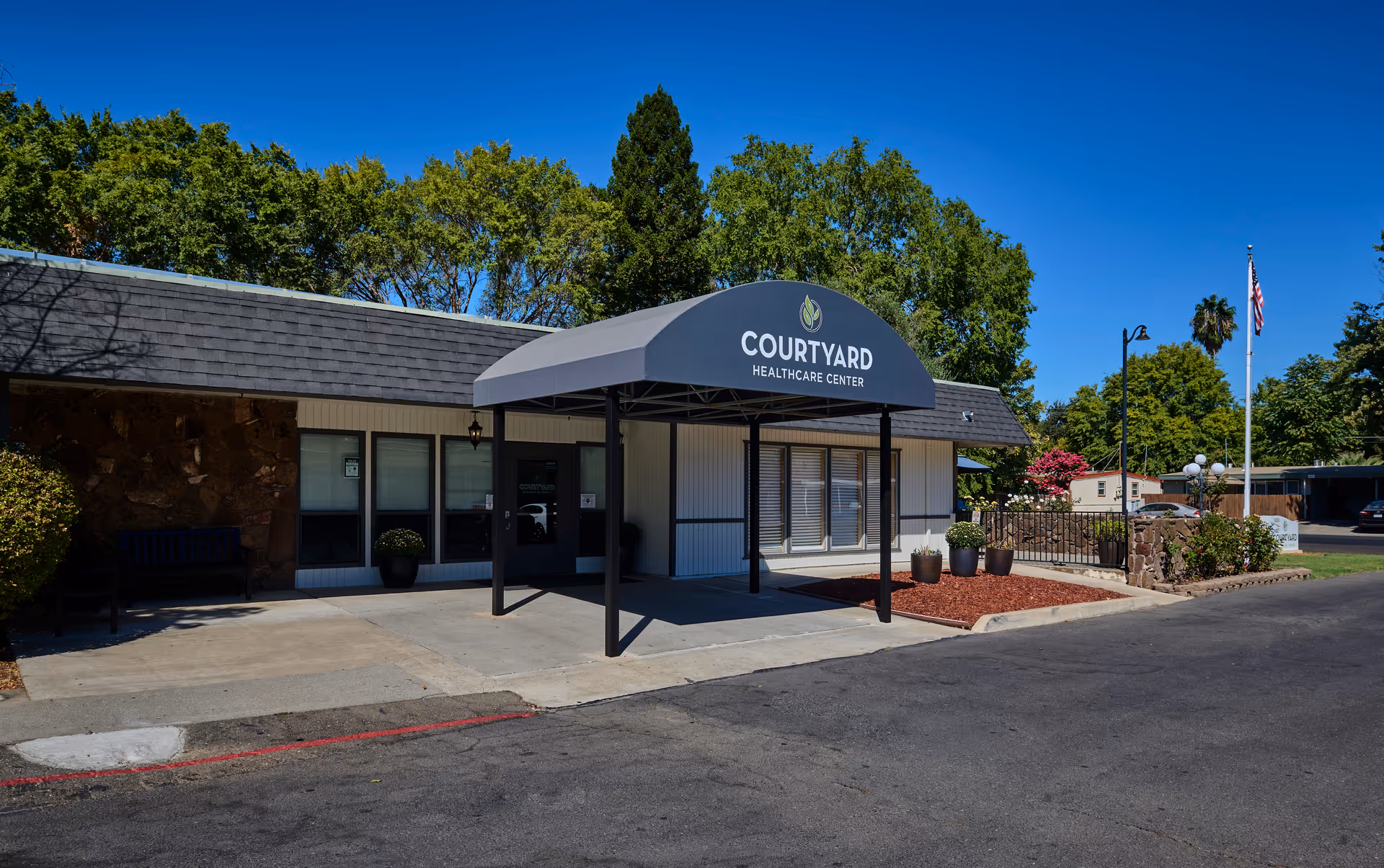 Exterior view of Courtyard Healthcare Center building entrance with a covered awning, potted plants, and a clear blue sky. There is a flagpole with an American flag and trees in the background.