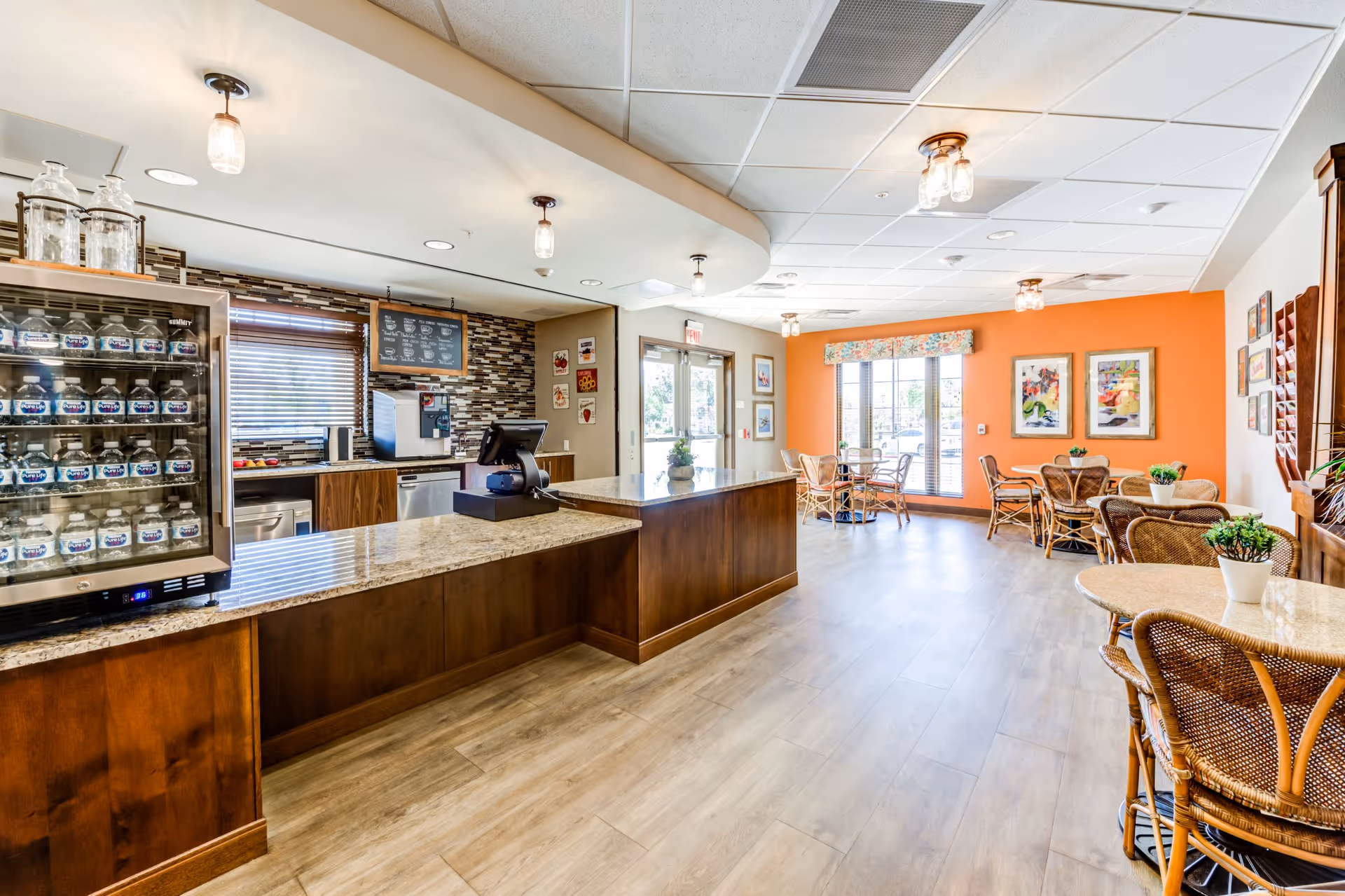 Bright and clean dining area with wooden tables and wicker chairs, a counter with a granite top, a refrigerated display case stocked with bottled water, and a coffee machine behind the counter. The room has large windows with blinds, orange accent wall with framed artwork, and light wood flooring.