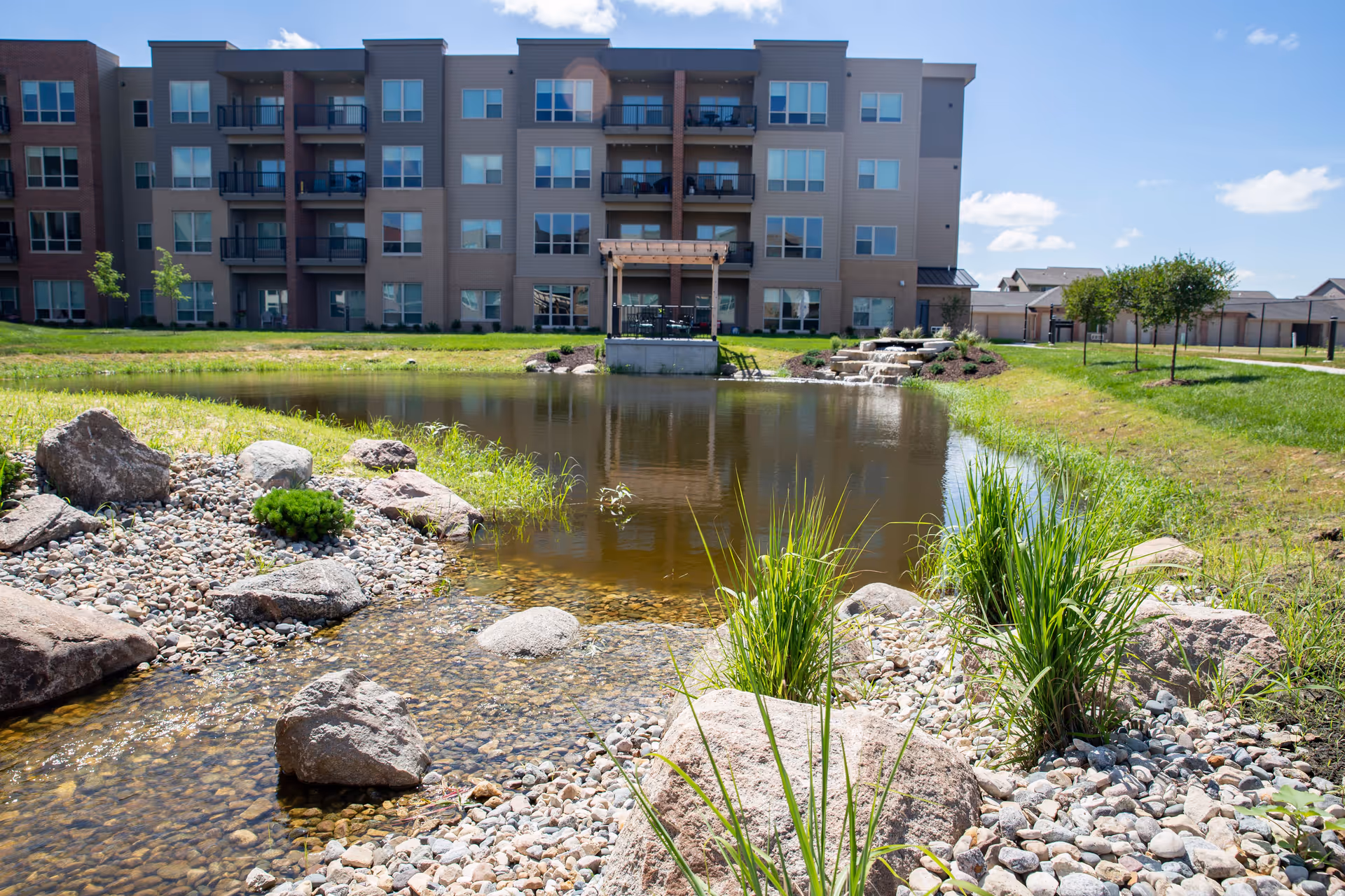 Apartment-style senior living building with balconies overlooking a landscaped pond, rocks, and greenery.