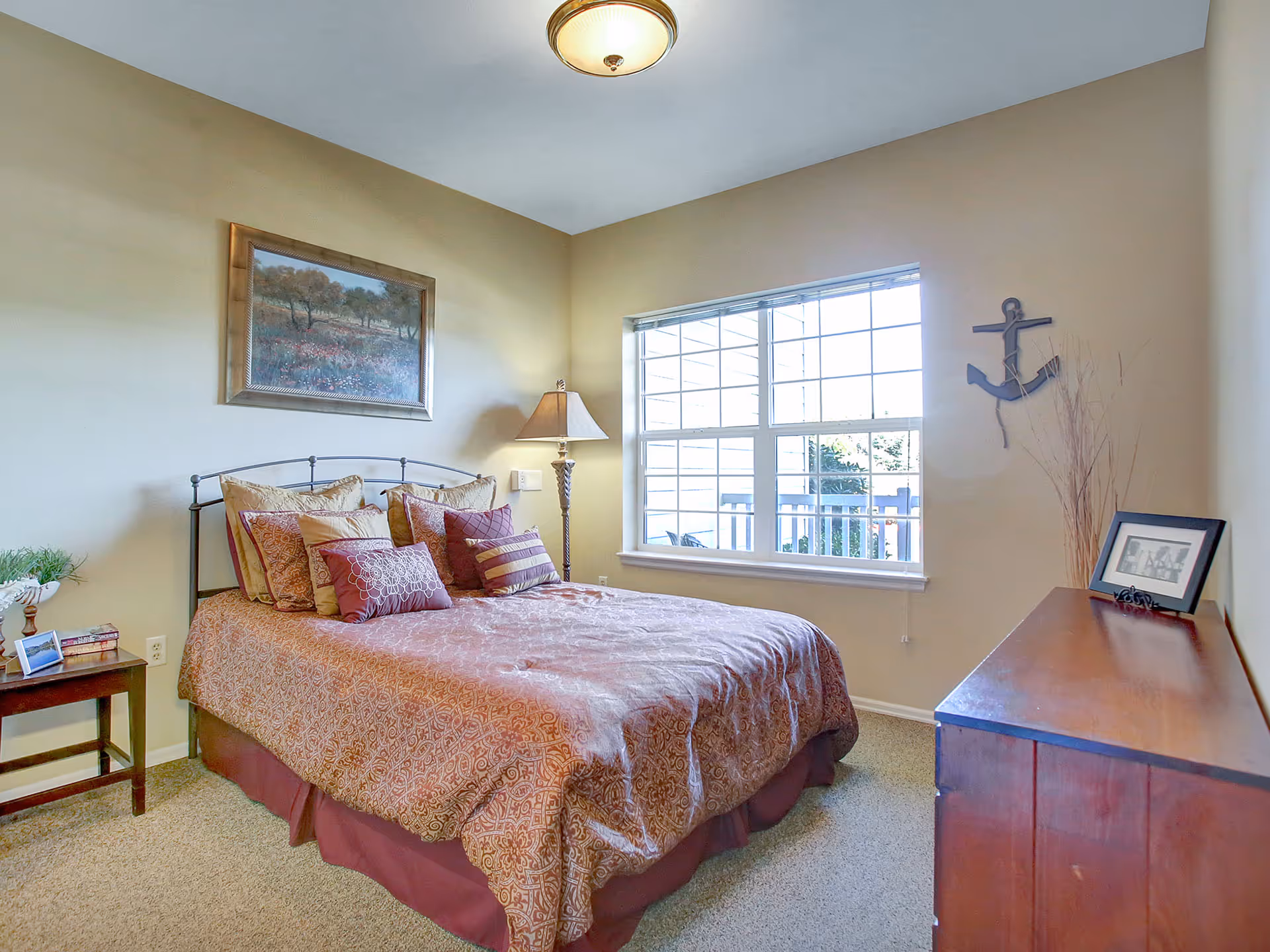 A cozy bedroom with a double bed covered in a patterned bedspread and multiple pillows. There is a wooden nightstand with a small plant, books, and a photo frame on the left side of the bed. A floor lamp stands next to the bed. A large window with white framing lets in natural light. On the right wall, there is a wooden dresser with a framed picture and decorative items, and a metal anchor wall decoration above it. The walls are painted beige and the carpet is light brown.