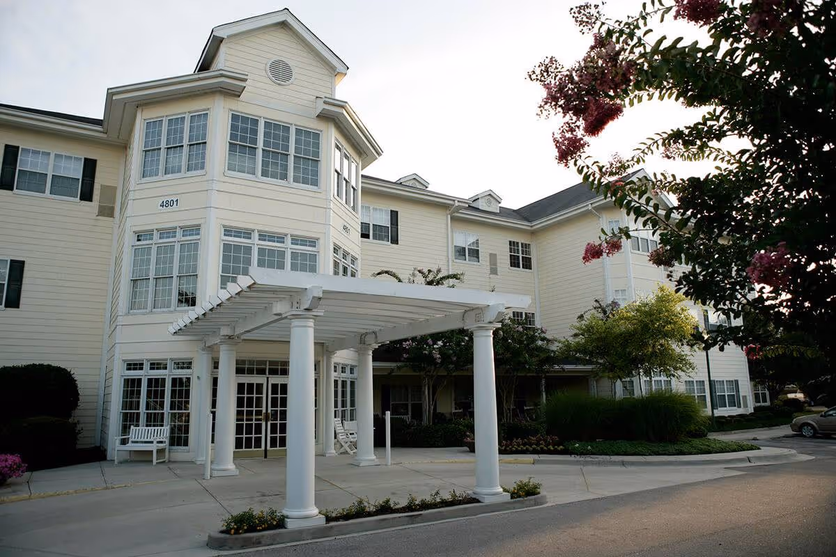 Exterior view of HarborChase Of Huntsville Specialty Care building featuring a three-story cream-colored structure with multiple windows, a white pergola supported by columns at the entrance, surrounding greenery, and a driveway with parked cars.