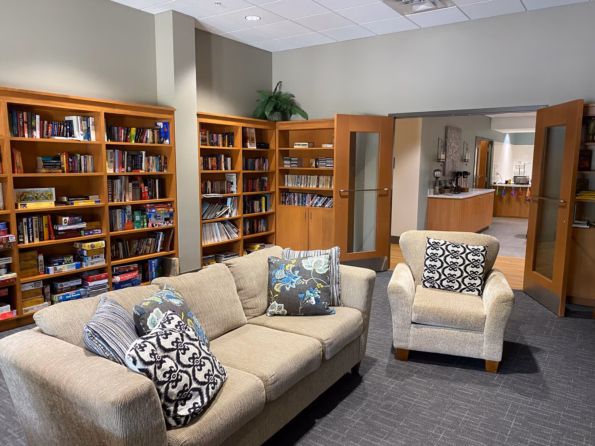 A cozy common room with a beige sofa and armchair, patterned pillows, wooden bookshelves along the wall, and double doors opening to a dining/serving area.