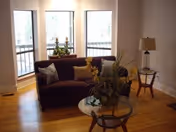 A cozy living room with a dark brown sofa adorned with decorative pillows, positioned near three large windows. In front of the sofa is a round glass coffee table with a floral arrangement. To the right, there is a wooden side table with a lamp and another small decorative item. The room has light-colored walls and wooden flooring.