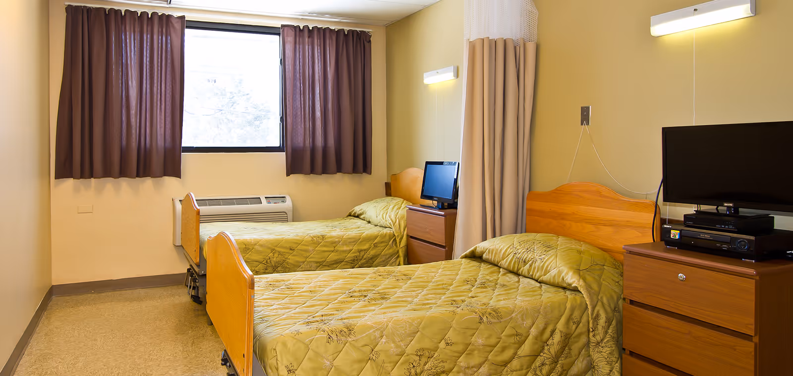 A small room with two single beds covered with green patterned bedspreads. Each bed has a wooden headboard and is positioned against opposite walls. Between the beds is a wooden nightstand with a small television and electronic devices on top. A window with dark curtains is on the far wall, and a beige privacy curtain hangs between the beds. The walls are painted light yellow, and the floor has a beige linoleum surface.