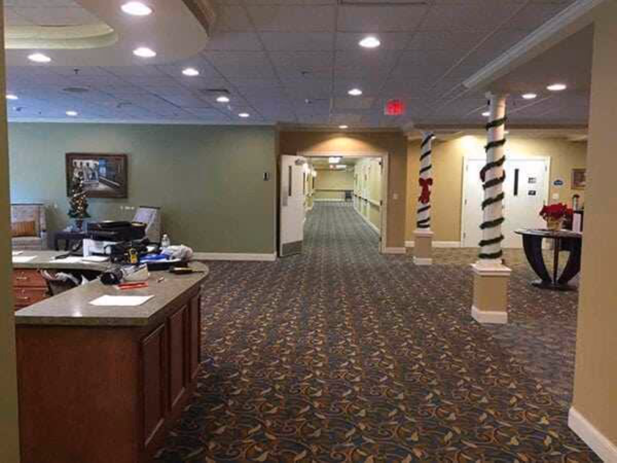 Interior view of a senior living facility hallway with patterned carpet, a reception desk on the left, two columns wrapped with garland and red bows, and a table with a poinsettia plant on the right. The hallway extends into the distance with doors and handrails along the walls.