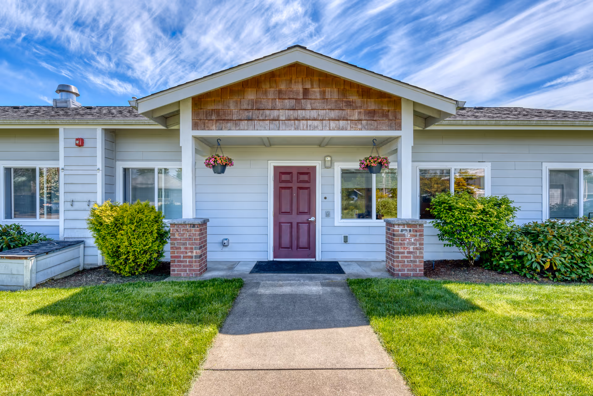 Front exterior view of a single-story building with a red door centered under a small covered porch supported by brick pillars. The building has light gray siding, white trim, and hanging flower pots on either side of the door. There is a concrete walkway leading up to the entrance, surrounded by well-maintained green grass and shrubs.