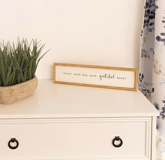 A white dresser with two black ring handles, a small rectangular wooden planter with green grass-like plants, and a framed sign that reads 'Start each day with grateful heart' placed on top. A floral curtain is partially visible on the right side.