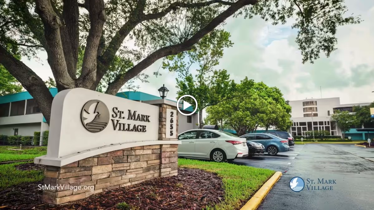 Entrance sign for St. Mark Village with a large tree behind it and a parking lot with several cars parked. The building of the facility is visible in the background under a cloudy sky.