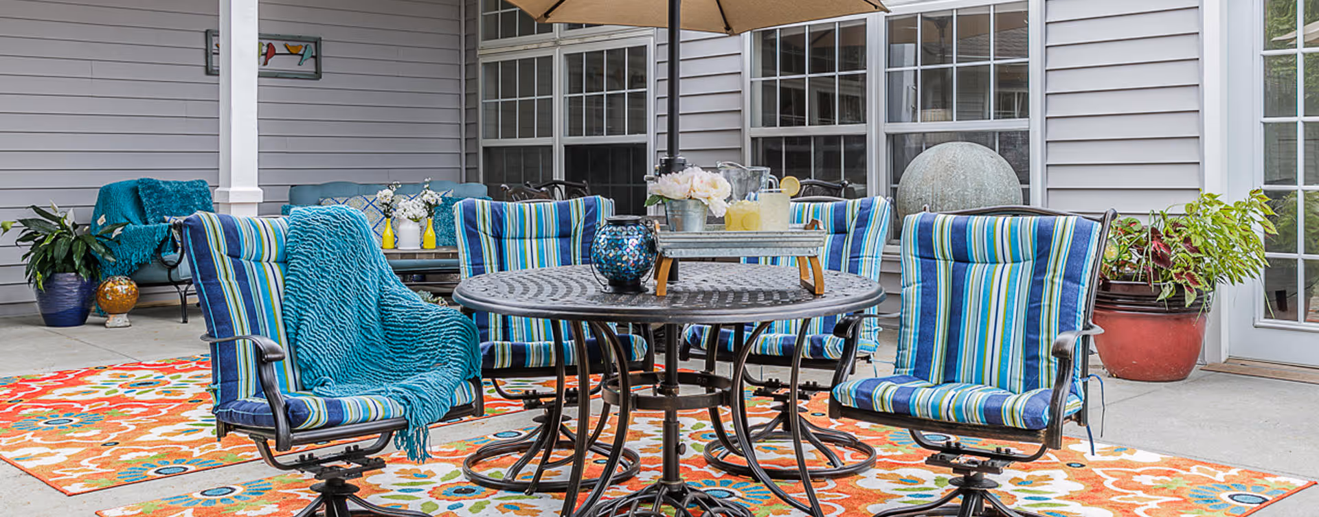 Outdoor patio area with a round metal table surrounded by six striped cushioned chairs. The table has a tray with a pitcher of lemonade and glasses. The patio is decorated with colorful rugs, potted plants, and a large umbrella providing shade. The background shows windows and siding of the building.