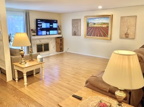 A cozy living room with light wood flooring, a beige couch, two wooden side tables each with a lamp, a wall-mounted flat screen TV above a white fireplace, and three framed artworks on the walls.