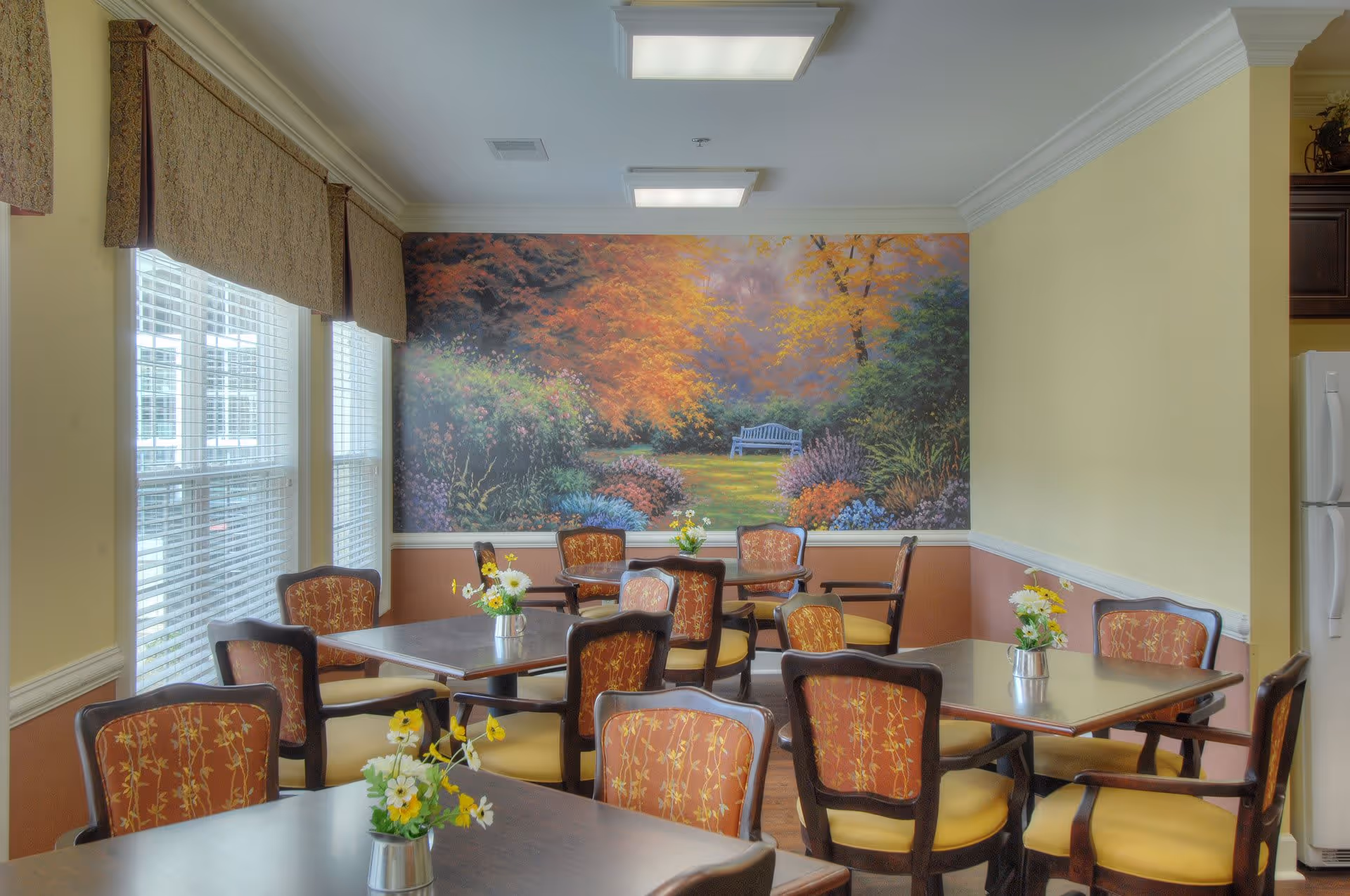 Dining room with several tables and upholstered chairs, small floral centerpieces, large windows on the left, and a colorful garden mural on the far wall.