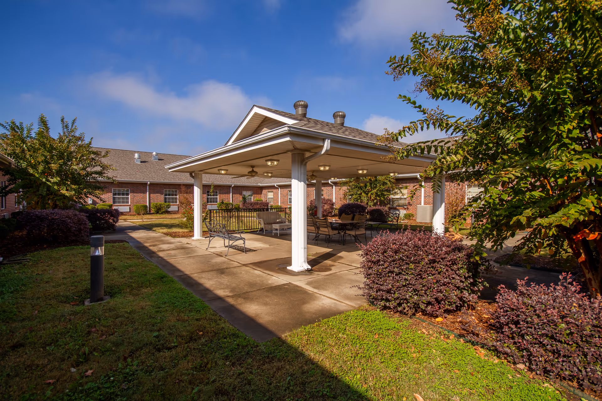 Covered outdoor pavilion with chairs and tables in a landscaped courtyard of a brick senior living facility under a blue sky.