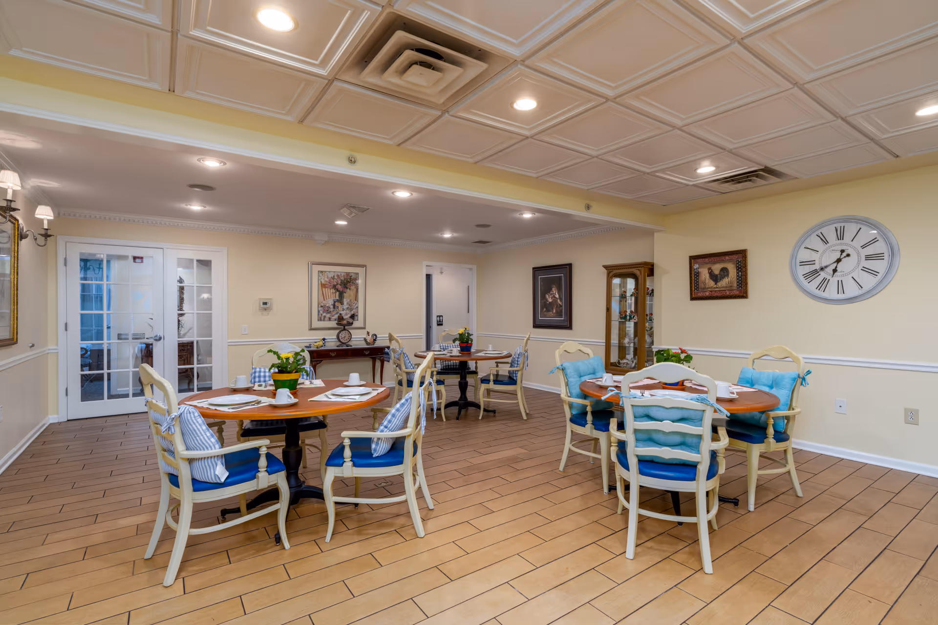 A cozy dining room with round wooden tables and cushioned chairs arranged neatly. The room has light yellow walls adorned with framed artwork and a large wall clock. The floor is tiled with a wood-like pattern, and there are double glass doors on one side. The tables are set with placemats, cups, and small potted plants as centerpieces.