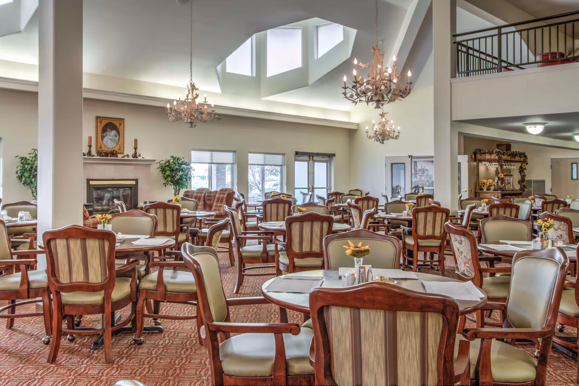 Spacious dining room with multiple round wooden tables and cushioned chairs arranged neatly. The room features a high ceiling with large skylights, two ornate chandeliers, a fireplace with a framed portrait above it, and large windows letting in natural light. There are plants and decorative elements around the room, creating a warm and inviting atmosphere.