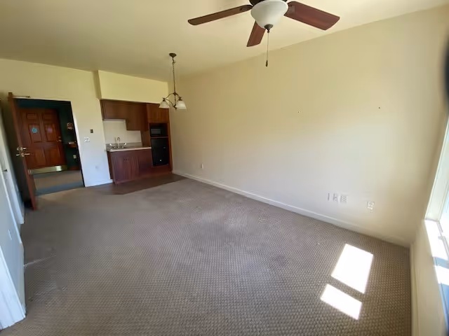 Empty room with beige carpet and cream-colored walls, featuring a ceiling fan with light, a small kitchen area with wooden cabinets and a sink, a hanging light fixture, and a window letting in sunlight. The entrance door is visible in the background.
