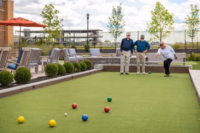 Three elderly people playing bocce ball on a green bocce court outdoors at The Summit of Blue Ash. There are chairs and tables with umbrellas along the side, some bushes, and a fence in the background under a partly cloudy sky.