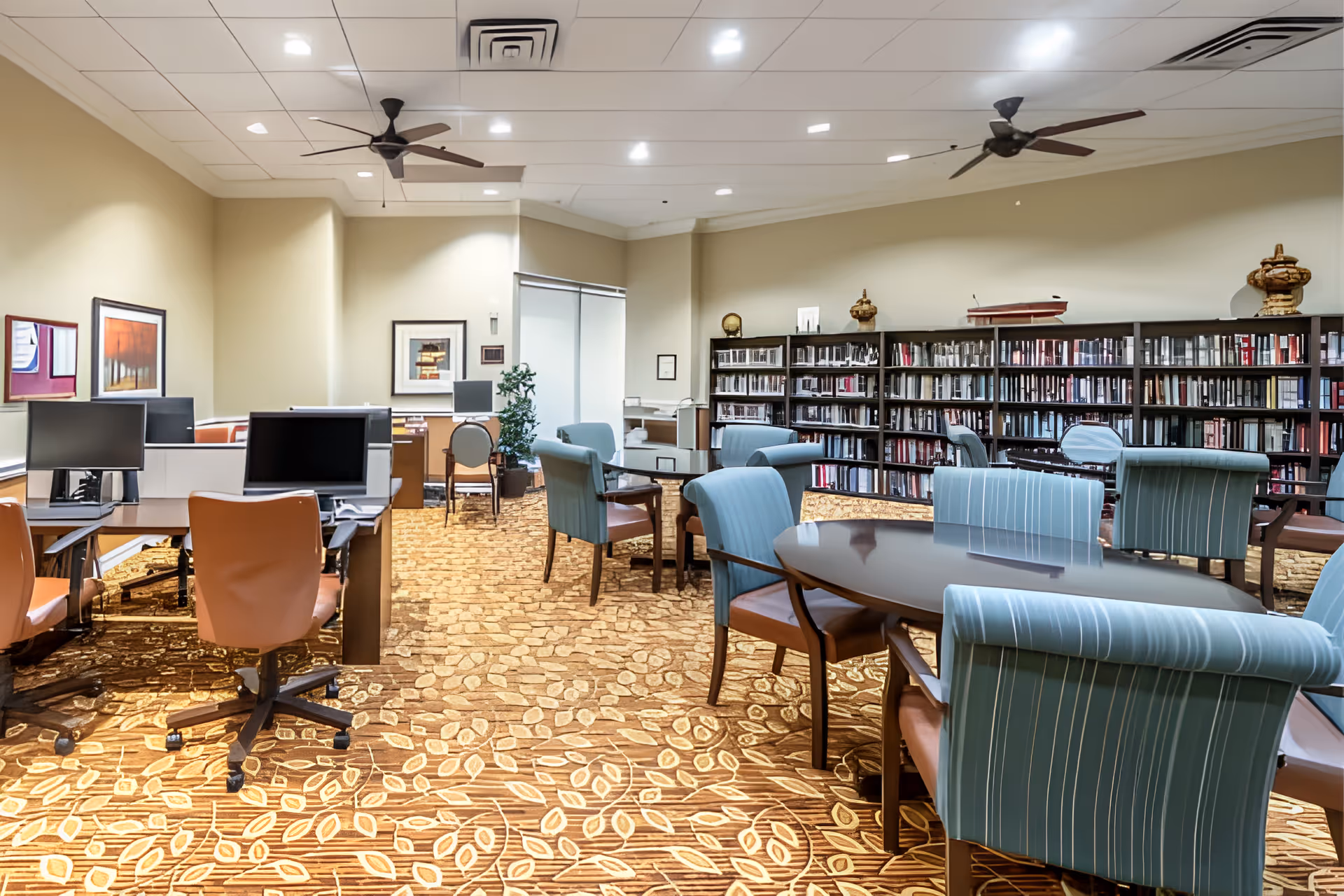 A spacious room with multiple seating areas including tables with blue cushioned chairs and desks with brown office chairs and computer monitors. The room has a patterned carpet, beige walls, ceiling fans, and a large bookshelf filled with books along one wall. There are framed pictures on the walls and a potted plant near the back.