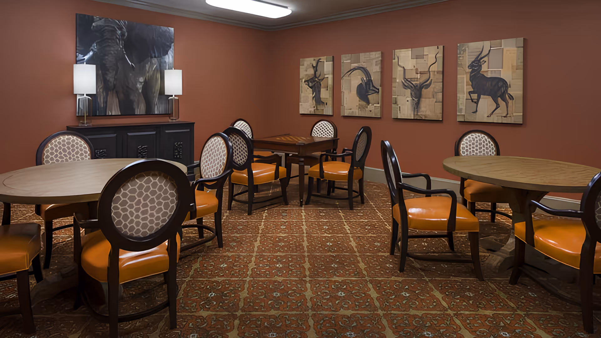 A communal dining room with round wooden tables, orange-upholstered chairs, patterned carpet and animal artwork on the walls.