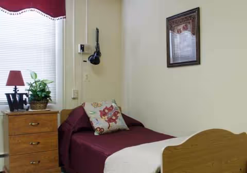 Single bedroom in a senior living facility with burgundy bedding, dresser, plant, and framed art.