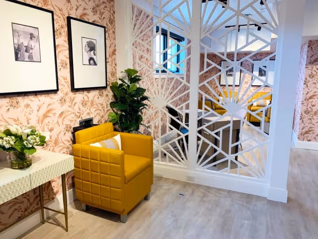 A cozy interior corner featuring a mustard yellow armchair with a white pillow, a white console table with a vase of flowers, two black and white framed pictures on a pink floral wallpapered wall, and a decorative white partition with geometric patterns separating the space.
