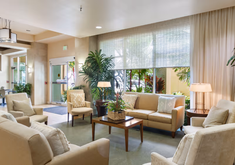 Well-lit lounge with beige sofas and armchairs arranged around a wooden coffee table in front of a large window and potted plants.