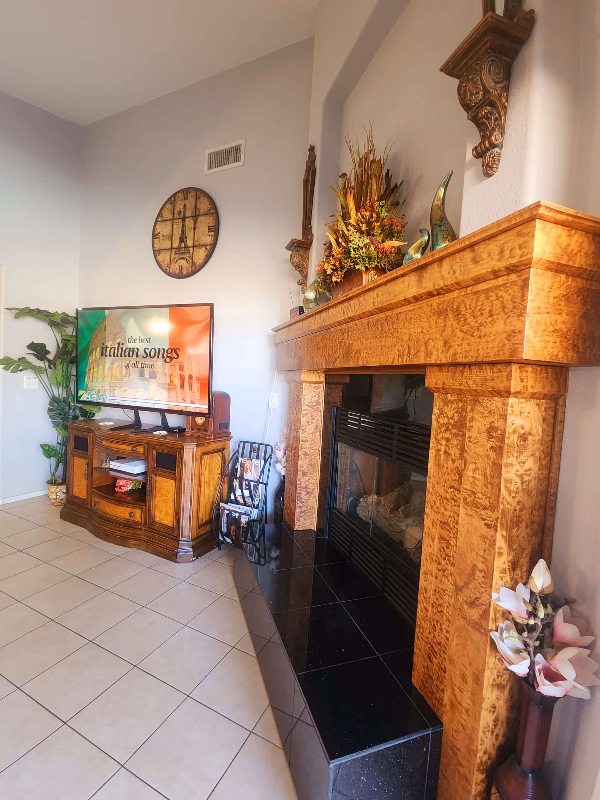A cozy living room corner featuring a wooden fireplace mantel decorated with a floral arrangement and decorative items. To the left, there is a wooden TV stand with a flat-screen TV displaying 'the best italian songs of all time' with an Italian flag background. A round wall clock with an Eiffel Tower design hangs above the TV. The floor is tiled, and a potted plant is visible in the corner.