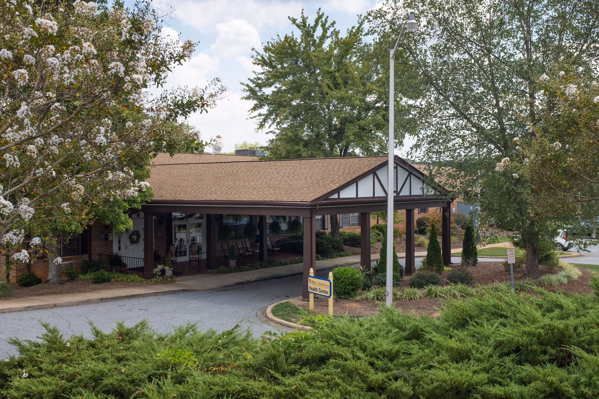 Entrance to a health center building with a covered driveway, surrounded by trees and shrubs under a partly cloudy sky.