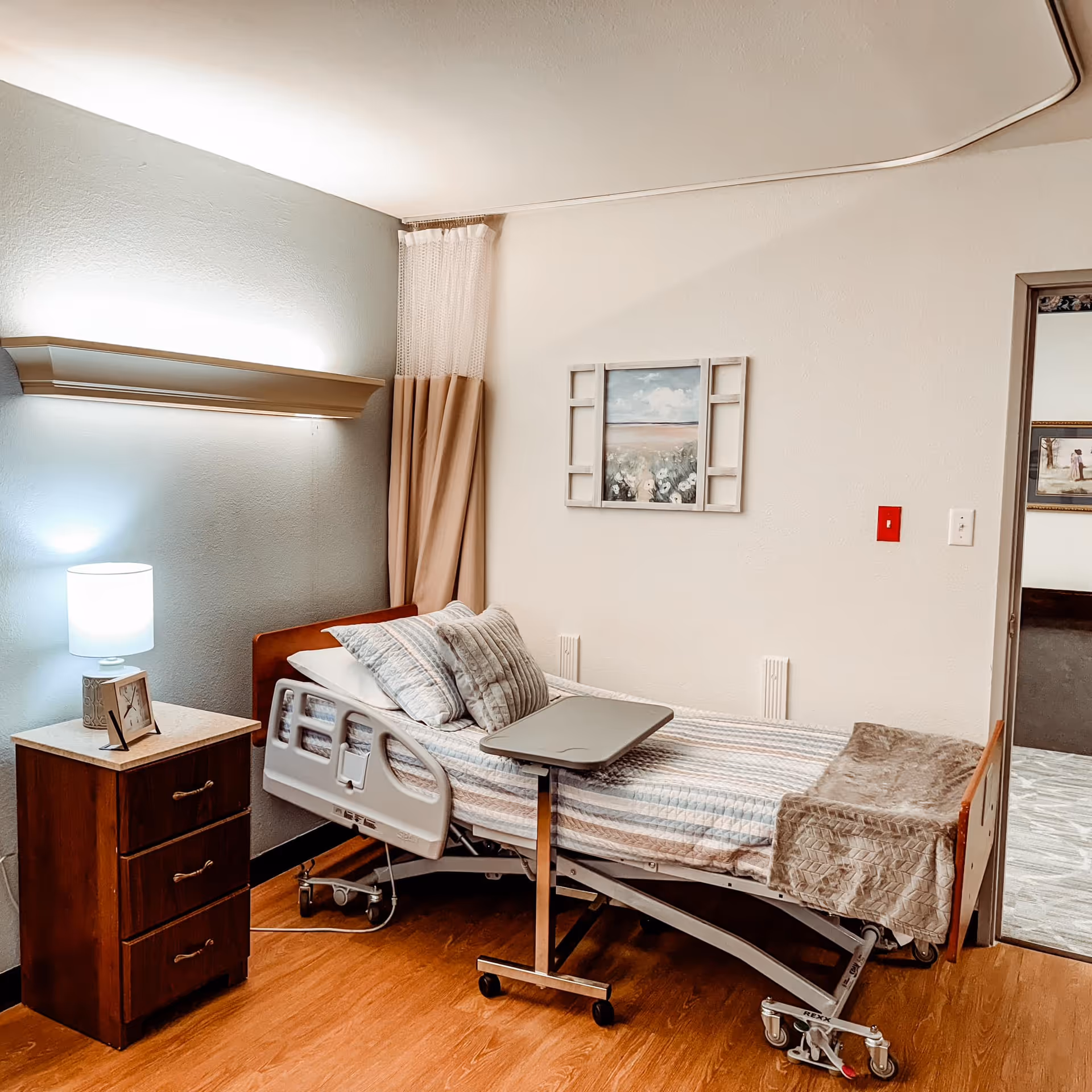A tidy patient bedroom with an adjustable hospital bed, overbed table, bedside dresser with lamp, wall art and curtains.