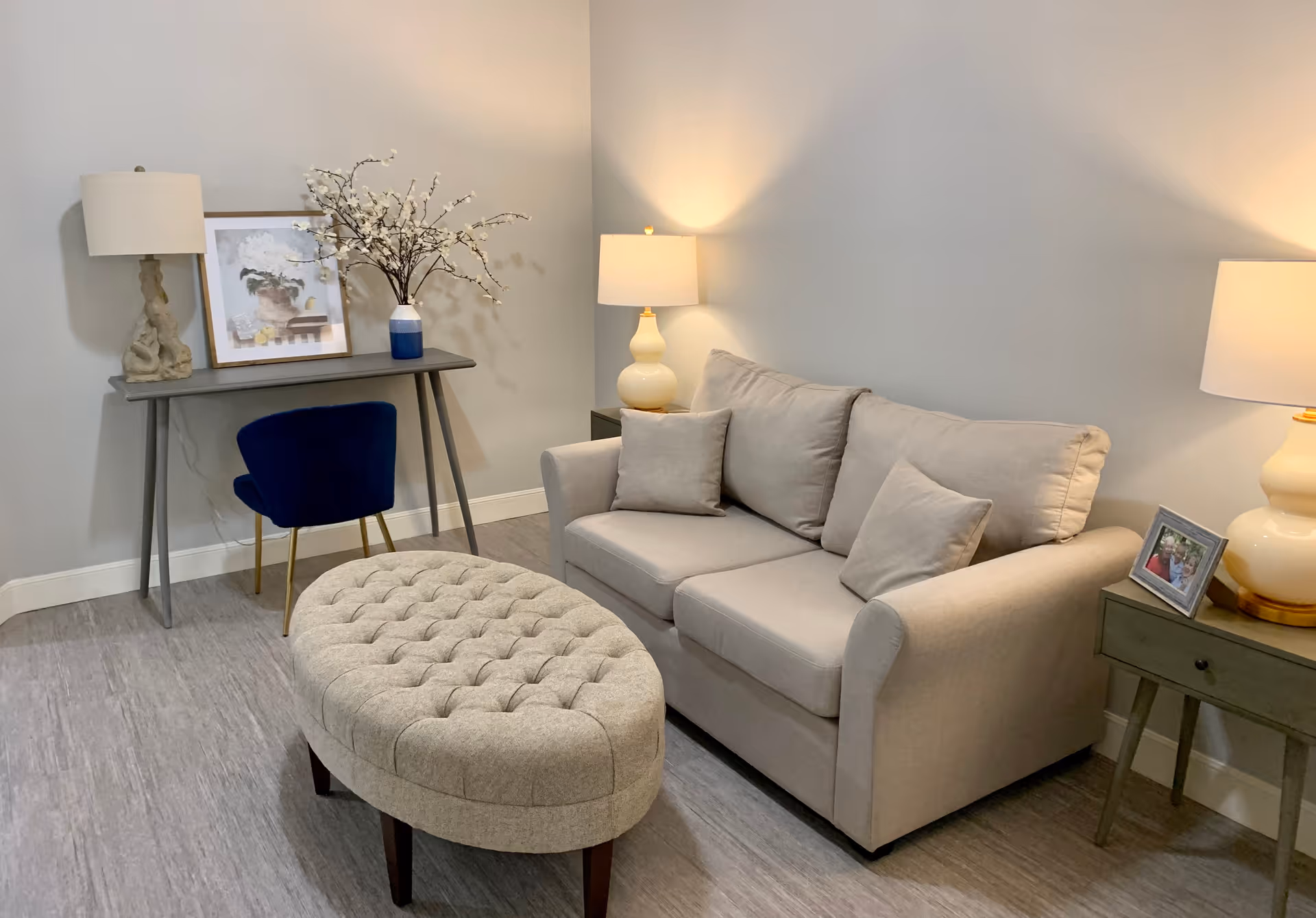 A cozy living room with a beige sofa adorned with three cushions, a tufted oval ottoman in front, two side tables each with a lamp, and a gray console table against the wall with a blue chair, a framed picture, and a vase with white flowers.