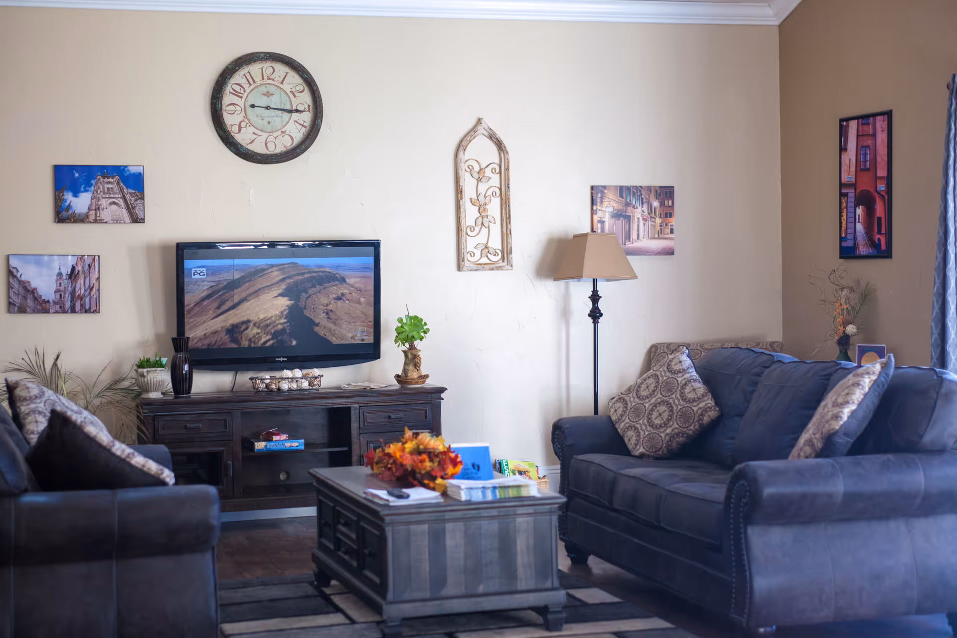 A cozy living room with two dark blue sofas adorned with patterned cushions, a wooden coffee table with a decorative centerpiece, a TV mounted on a wooden console, a floor lamp, and several framed pictures on the walls. A large clock is hung above the TV.