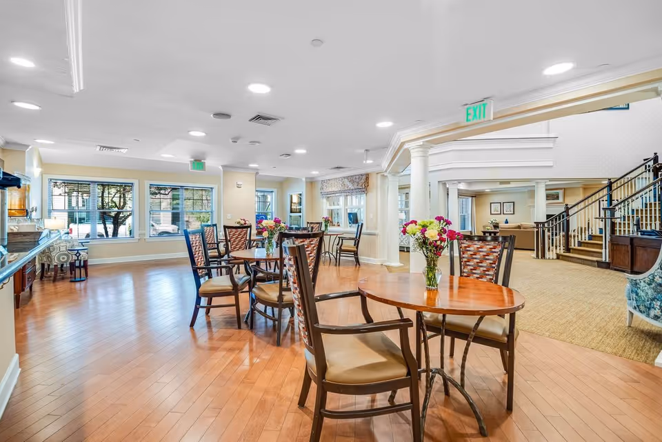 A bright and spacious common area in a senior living facility featuring wooden round tables with chairs arranged around them. Each table has a vase with colorful flowers. The room has large windows letting in natural light, light-colored walls, and a polished wooden floor. In the background, there is a carpeted area with a staircase and additional seating.