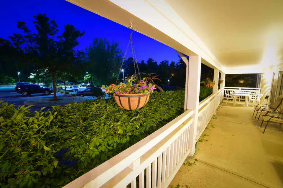 A well-lit covered porch at night with white railings and columns, hanging flower baskets, patio chairs, and a white table with benches. Beyond the porch, there are green bushes and trees, and a parking lot with several parked cars under streetlights.