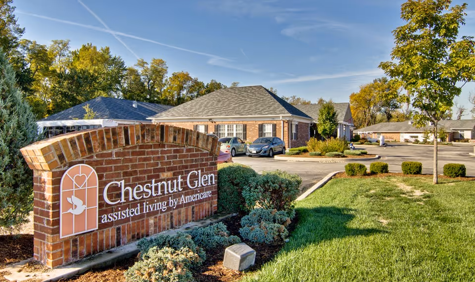 Exterior view of Chestnut Glen Senior Living facility showing a brick sign with the facility name and logo, surrounded by landscaped bushes and grass, with buildings and parked cars in the background under a clear blue sky.