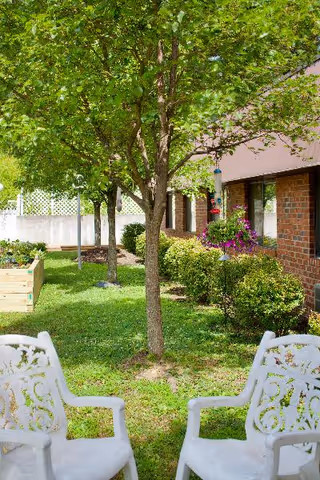 Outdoor garden area at a senior living community featuring two white decorative chairs facing a tree with green leaves. There are bushes, a hanging flower basket, and a raised garden bed near a brick building with windows.