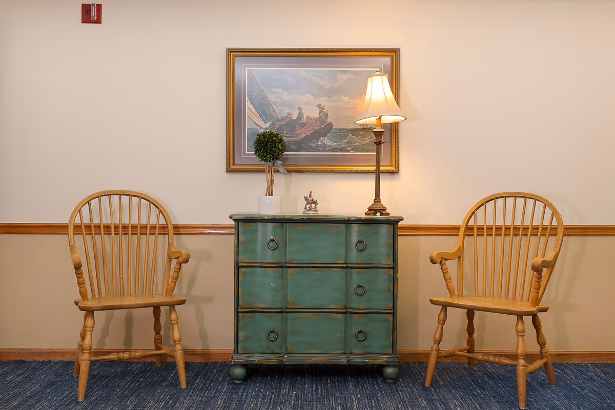 Two wooden armchairs flank a distressed green chest of drawers with a small potted plant and a table lamp on top. Above the chest hangs a framed painting of people sailing on a boat. The room has beige walls with a wooden chair rail and blue carpeted floor.