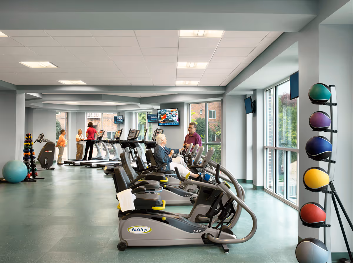 Bright fitness room with treadmills and NuStep exercise machines, medicine balls, and a few residents using equipment by large windows.
