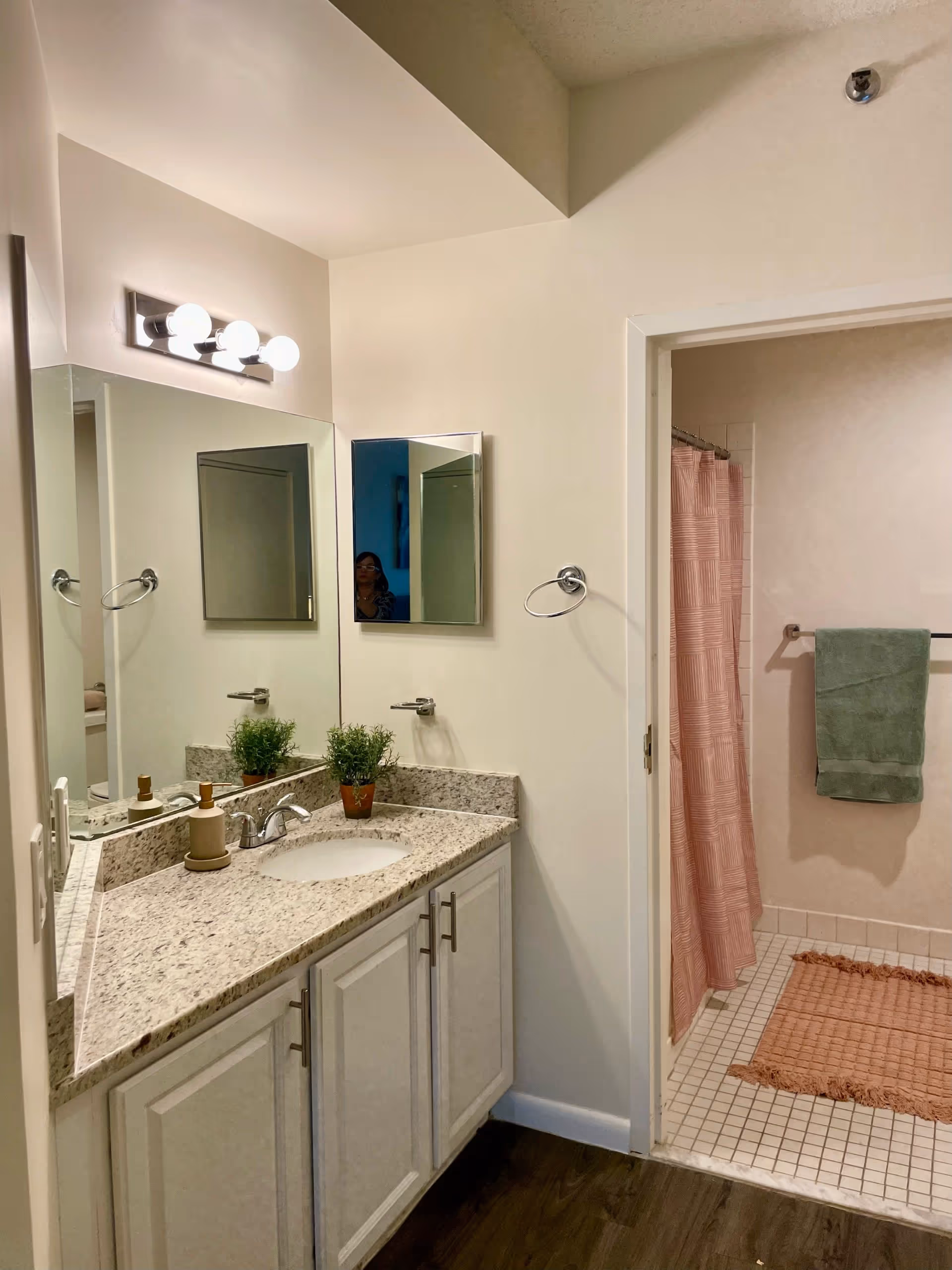 A bathroom with a granite countertop vanity featuring a sink, soap dispenser, and two small potted plants. Above the vanity is a large mirror with three round light bulbs mounted on the wall. To the right, there is an open doorway leading to a shower area with a pink shower curtain, a green towel hanging on a towel rack, and a pink bath mat on the tiled floor.