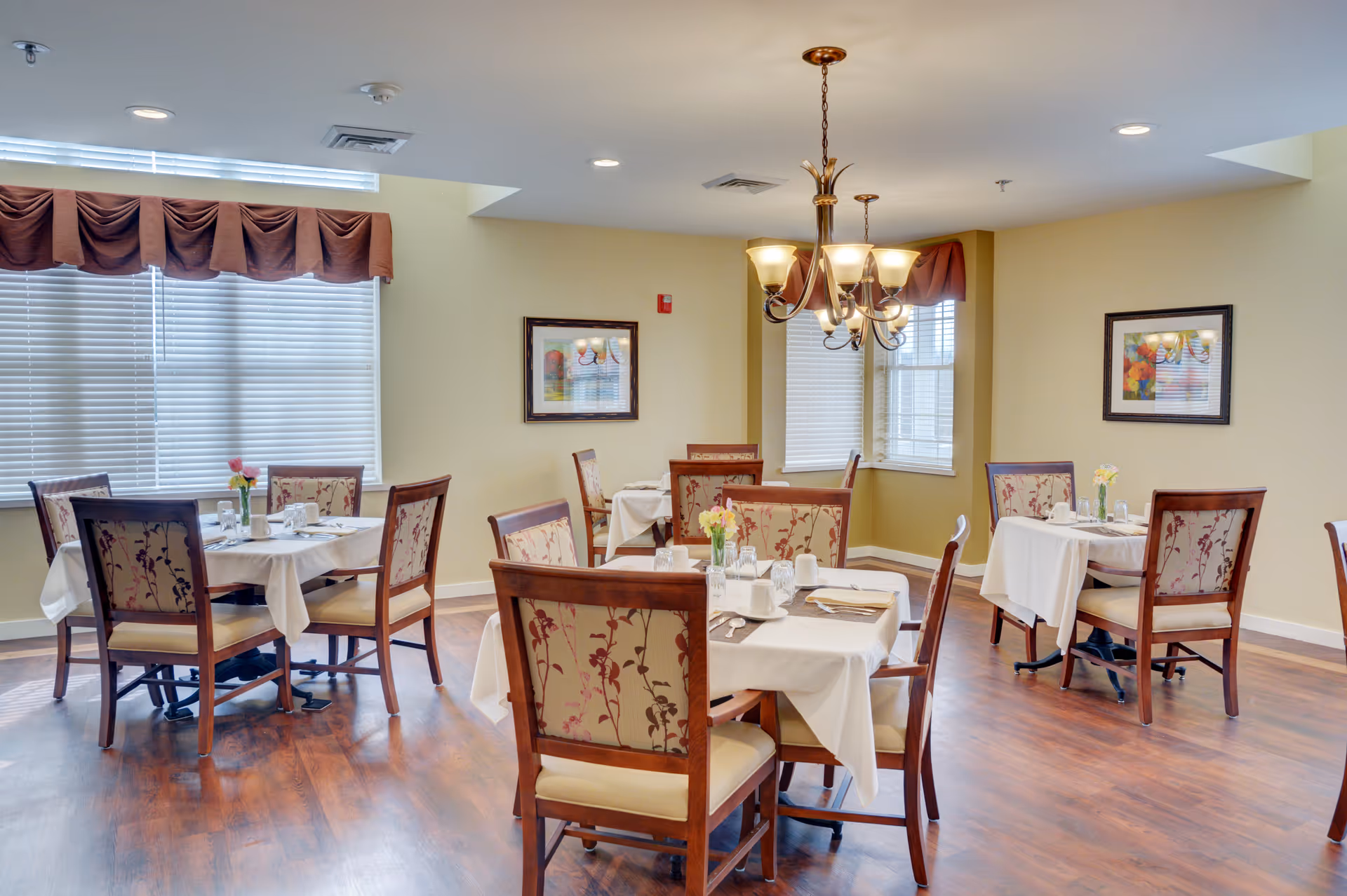 Bright dining room with multiple set tables, floral-upholstered chairs, a chandelier, and large windows with blinds.