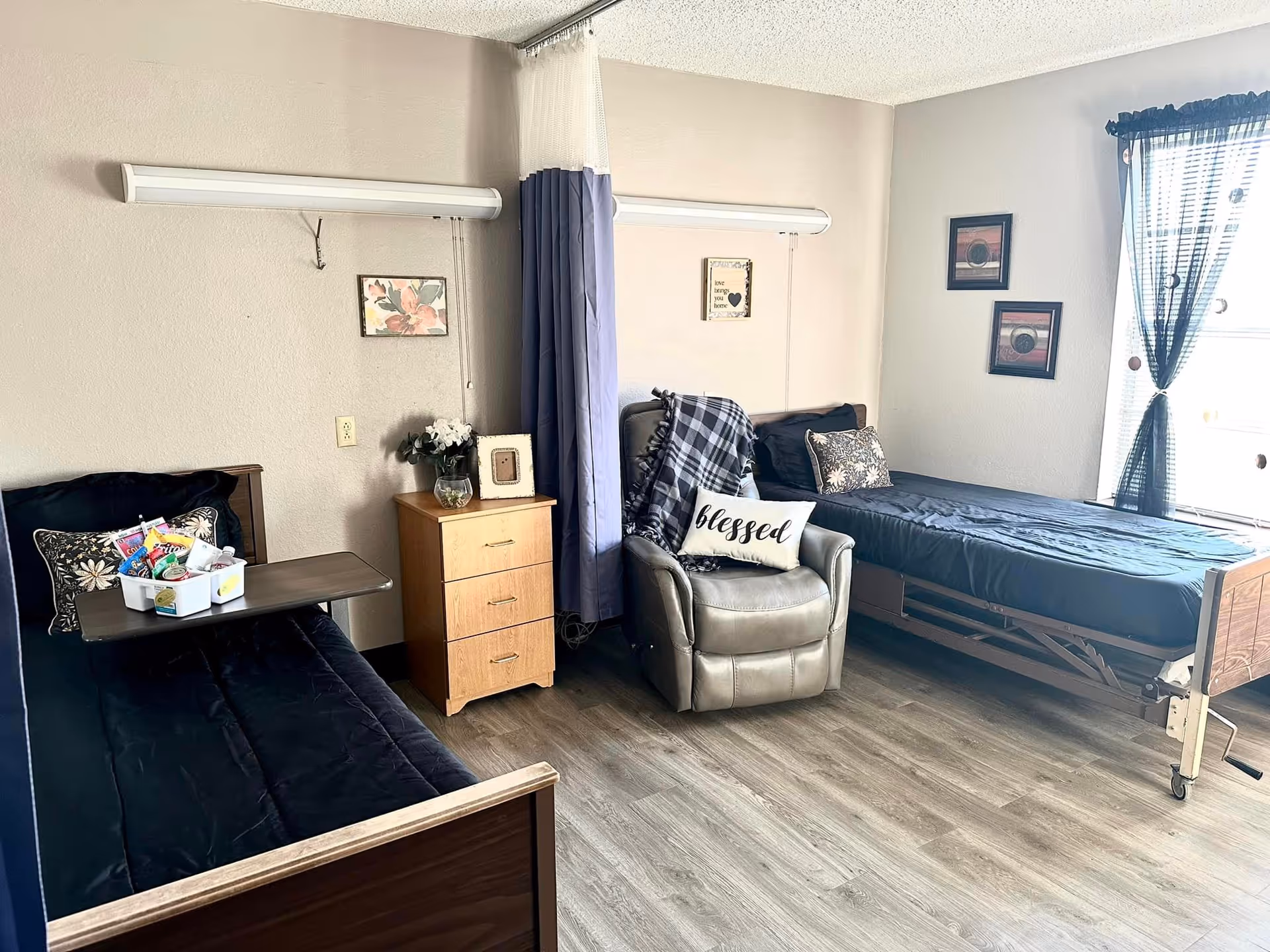 A room with two single beds, one on the left with a dark blue blanket and a tray with snacks on it, and one on the right with a dark blue blanket near a window with sheer curtains. Between the beds is a small wooden nightstand with a vase of flowers and a framed photo. In front of the nightstand is a gray recliner chair with a plaid blanket and a pillow that says 'blessed'. The walls are light-colored with framed pictures hanging, and the floor is wood laminate.
