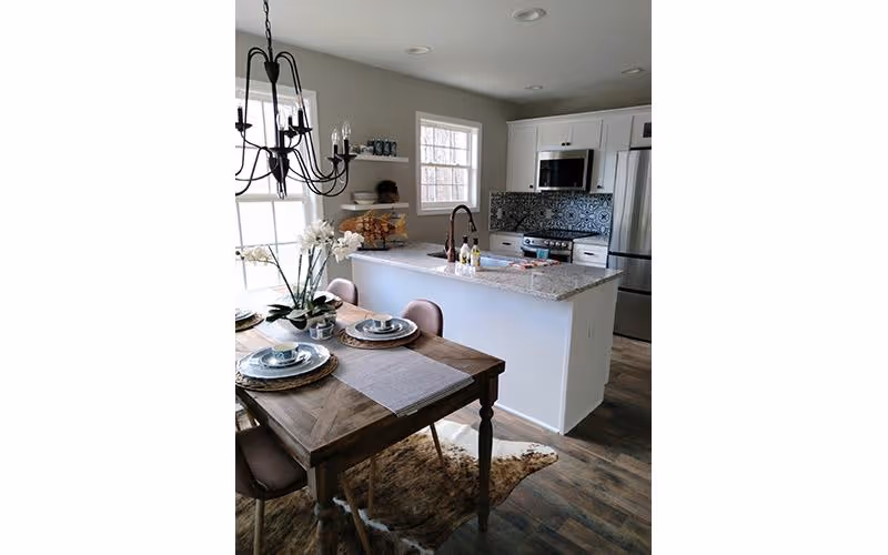 A modern kitchen and dining area featuring a wooden dining table set with plates and a flower centerpiece. The kitchen has a white island with a granite countertop, a sink with a bronze faucet, stainless steel appliances including a refrigerator and microwave, and a decorative tile backsplash. A black chandelier hangs above the dining table, and the floor is wood with a small animal hide rug underneath the table.