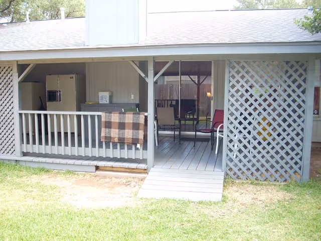 A covered outdoor porch area with a wooden floor and railing. There is a plaid blanket hanging on the railing, several chairs including a red and a beige one, and a small table. The porch has lattice panels on the right side and a ramp leading down to a grassy lawn.