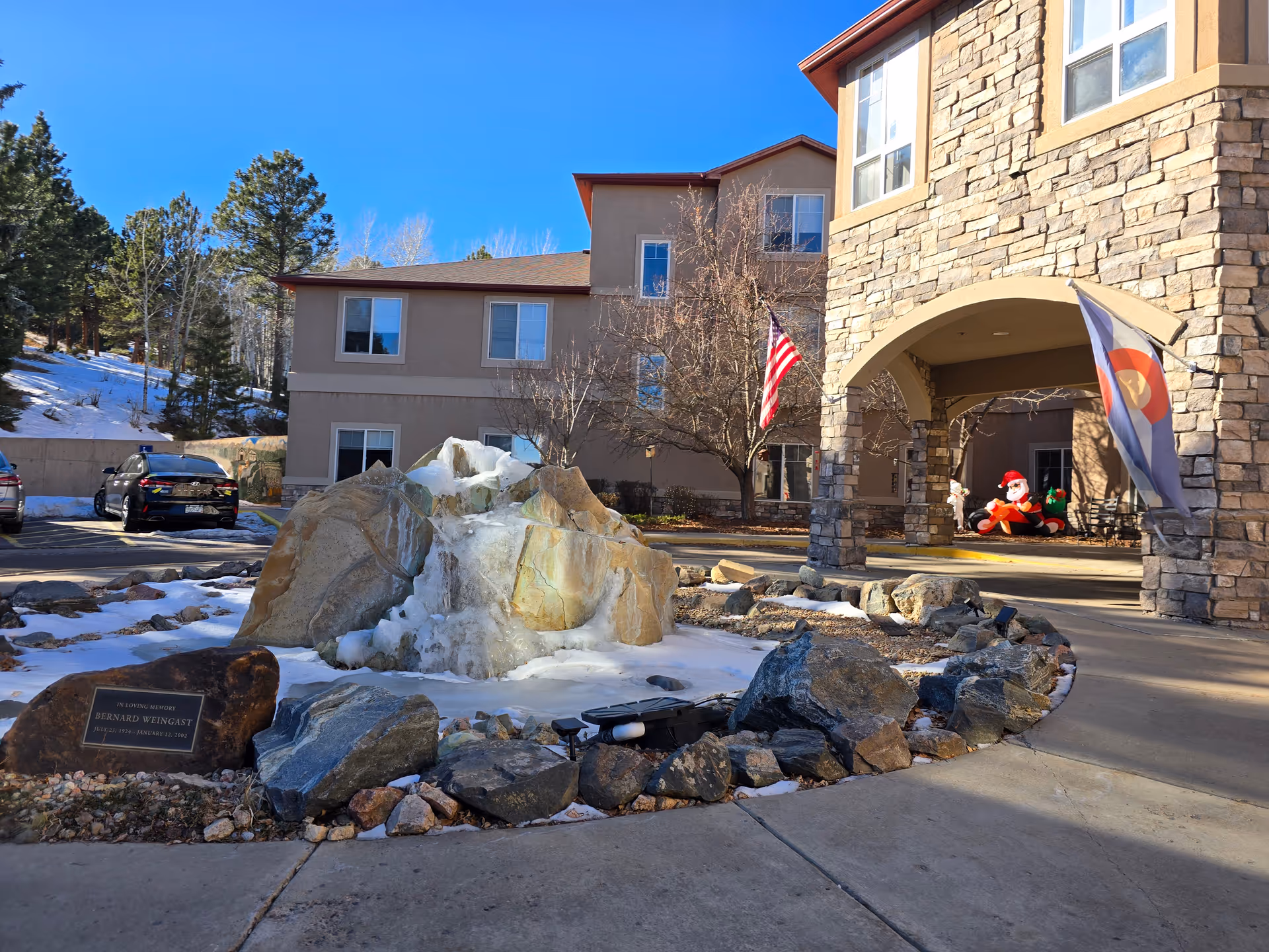 Outdoor view of Elk Run Assisted Living facility showing a stone and stucco building with multiple windows, an American flag and a Colorado state flag near the entrance. In front of the building is a circular rock garden with a frozen water feature and a memorial plaque. There are a few parked cars and some trees with snow on the ground under a clear blue sky.