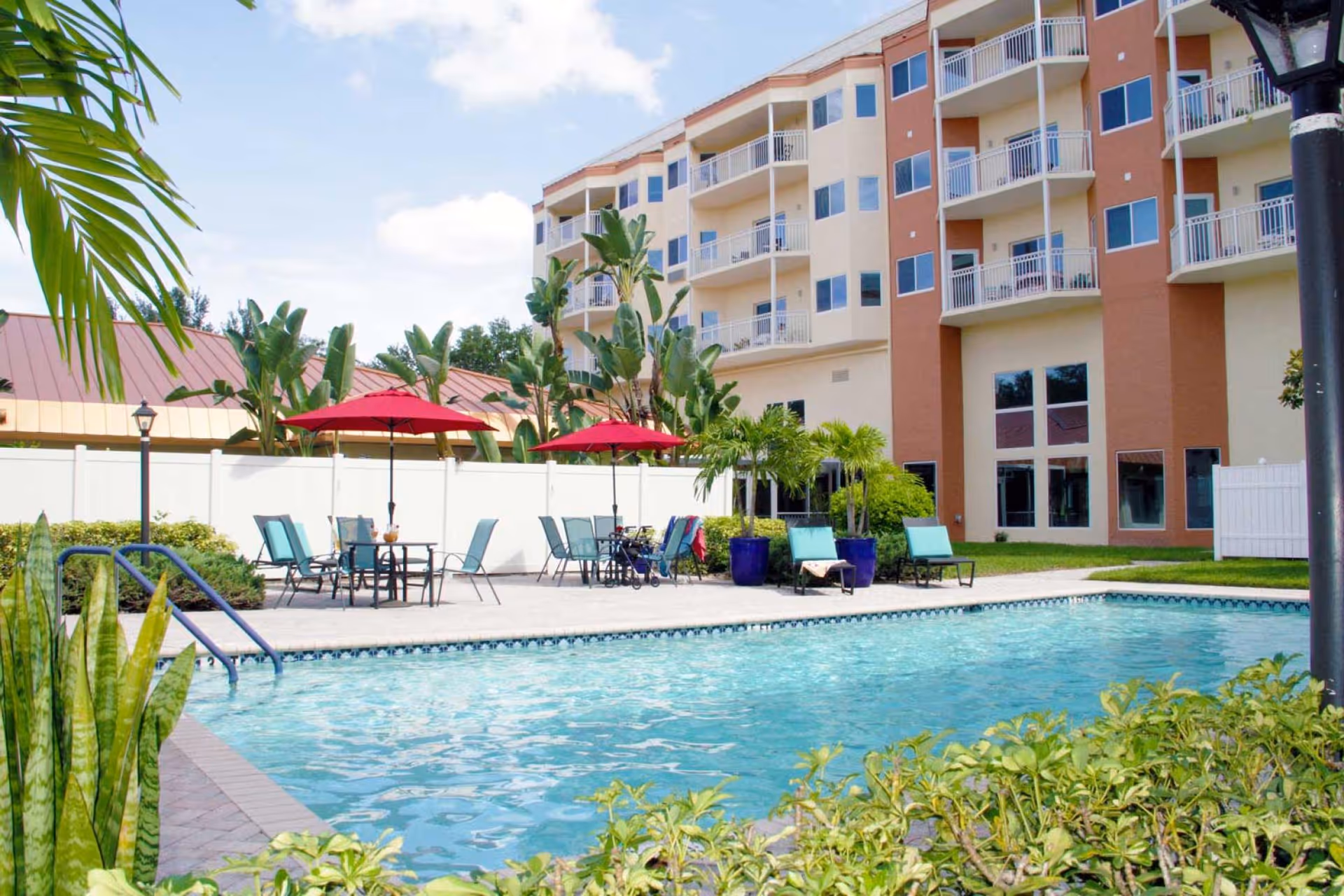Outdoor swimming pool area with clear blue water surrounded by lounge chairs and tables with red umbrellas. The background shows a multi-story building with balconies and tropical plants around the pool area under a partly cloudy sky.