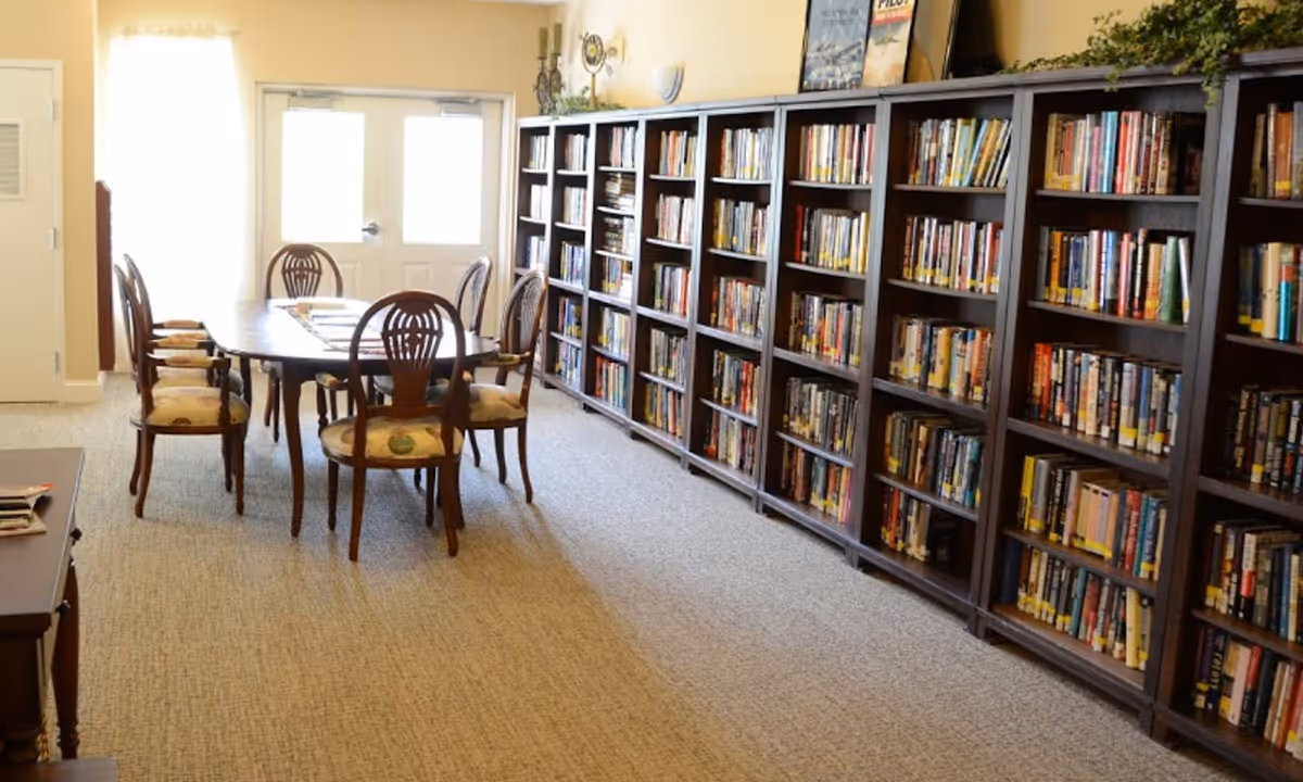 A bright room with a long wooden table surrounded by six wooden chairs with cushioned seats. Along the right wall, there are multiple tall bookshelves filled with books. The room has beige walls and carpeted flooring, with a double door at the far end letting in natural light.