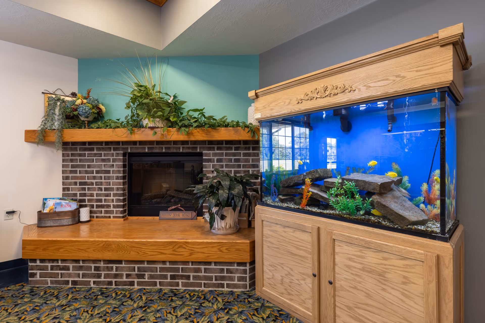 A cozy corner of a senior living facility featuring a brick fireplace with a wooden mantel decorated with green plants and flowers. Next to the fireplace is a large wooden aquarium filled with colorful fish, rocks, and aquatic plants. The room has a patterned carpet and walls painted in light colors with a teal accent behind the fireplace.