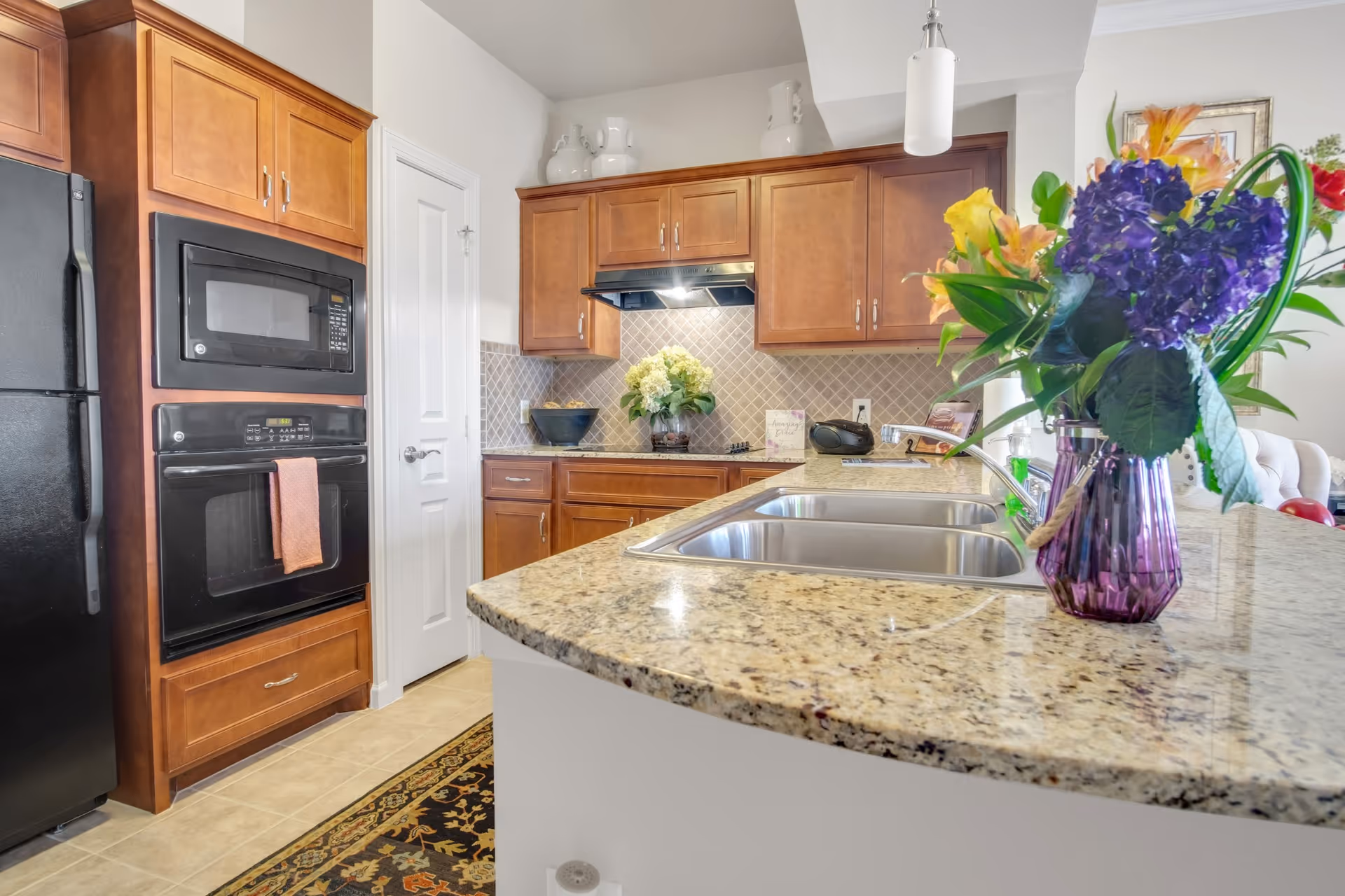 A modern kitchen with wooden cabinets, a black refrigerator, built-in microwave and oven, granite countertops, a double sink, and a vase with colorful flowers on the counter. The kitchen has a tiled backsplash and a small rug on the floor.