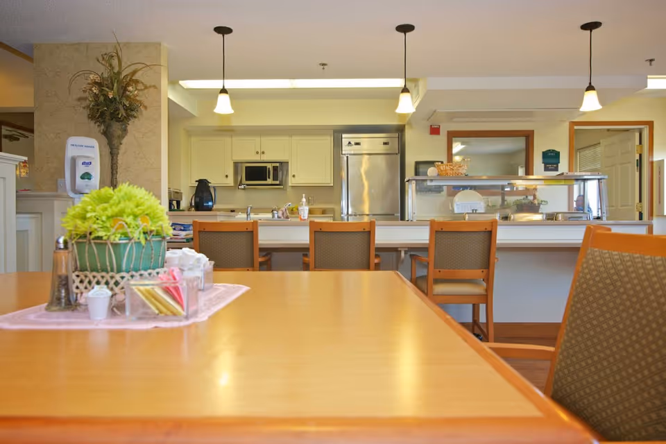 Interior view of a senior living facility dining area with a wooden table in the foreground decorated with a green potted plant and condiments. In the background, there is a kitchen area with white cabinets, a microwave, a stainless steel refrigerator, and a counter with chairs. Pendant lights hang from the ceiling, and a hand sanitizer dispenser is mounted on a wall to the left.