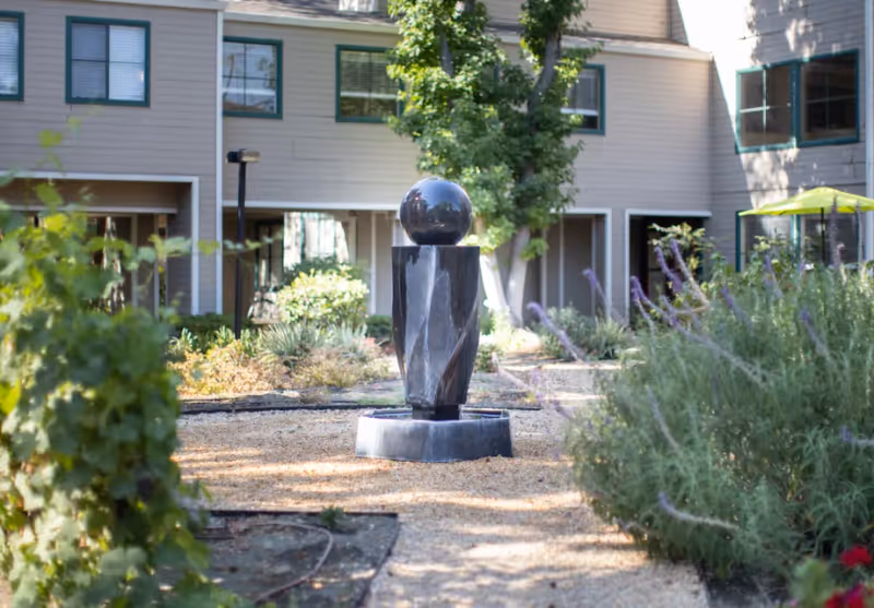 Outdoor garden area with a modern black stone sculpture featuring a spherical top, surrounded by plants and shrubs, with a multi-story residential building in the background.