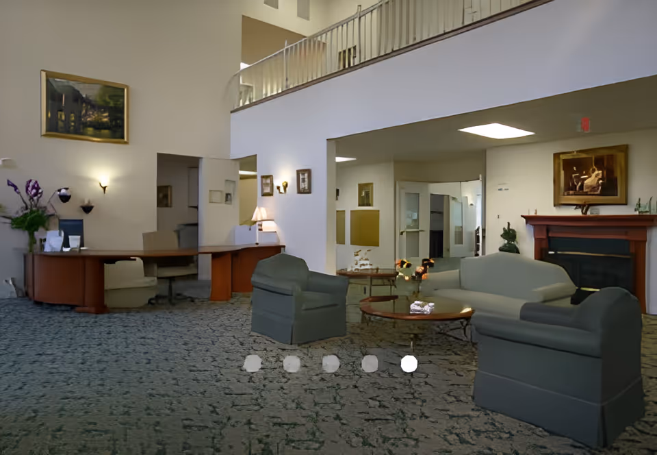 A spacious senior living facility lobby with a reception desk on the left, comfortable seating including armchairs and a sofa around a coffee table in the center, a fireplace with a painting above it on the right, and a carpeted floor. The walls are decorated with framed pictures and there is a balcony railing visible on the upper level.